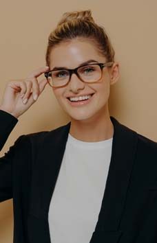 Positive happy young woman touching rim of glasses and looking through them, smiling female student touching spectacles while looking at camera with smile, isolated over beige wall with copy space